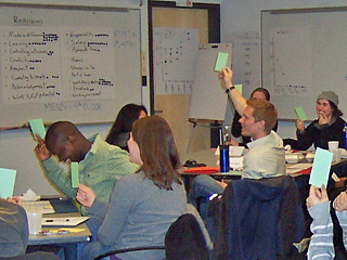 Students sitting in class raise their hands while holding green notecards. Students sitting in class raise their hands while holding green notecards.