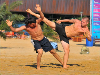 Two men engaging in capoeira martial arts on a beach. The man on the right is performing a high kick, and the man on the left has his arms extended upward and is using his foot to trip his opponent. Two men engaging in capoeira martial arts on a beach. The man on the right is performing a high kick, and the man on the left has his arms extended upward and is using his foot to trip his opponent.
