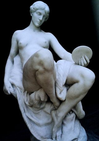 A marble statue of naked woman seated on a pedestal, legs crossed at the ankles, with linen draped between her thighs. A marble statue of naked woman seated on a pedestal, legs crossed at the ankles, with linen draped between her thighs.