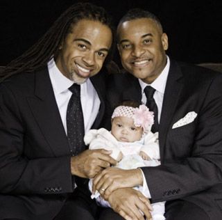 Two handsome, well-dressed men of color hold an infant, with a pink bow around her head. Two handsome, well-dressed men of color hold an infant, with a pink bow around her head.