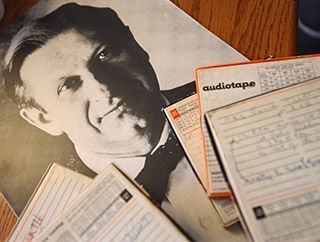 On a wooden tabletop, a black and white photograph of a distinguished-looking man wearing a bow tie rests beneath a pile of reel to reel tape boxes. On a wooden tabletop, a black and white photograph of a distinguished-looking man wearing a bow tie rests beneath a pile of reel to reel tape boxes.