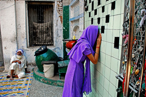 A photo of a girl looking through a window in Ahmedabad. Behind her is an old man, sitting on the ground, and a plastic bucket. By her right arm is a grating covered in padlocks. A photo of a girl looking through a window in Ahmedabad. Behind her is an old man, sitting on the ground, and a plastic bucket. By her right arm is a grating covered in padlocks.