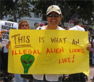 Photo of a woman in a baseball cap holding a bright yellow sign reading "THIS is what an illegal alien looks like!", with a sketch of a green bug-eyed alien next to a flying saucer. Photo of a woman in a baseball cap holding a bright yellow sign reading "THIS is what an illegal alien looks like!", with a sketch of a green bug-eyed alien next to a flying saucer.