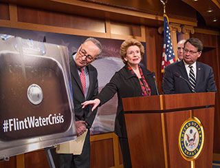 A red-haired, casually dressed woman, flanked by two suit-wearing gentlemen, stands at a podium and points to a large photo of a sink filled with dirty water and words reading: #FlintWaterCrisis. A red-haired, casually dressed woman, flanked by two suit-wearing gentlemen, stands at a podium and points to a large photo of a sink filled with dirty water and words reading: #FlintWaterCrisis.