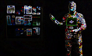 A human form stands in darkness, wearing a bodysuit illuminated from head to toe with various multi-colored circuits. A human form stands in darkness, wearing a bodysuit illuminated from head to toe with various multi-colored circuits.