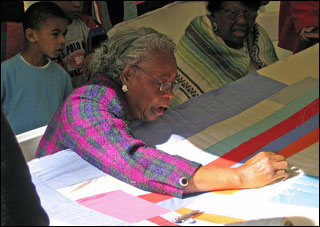 An elderly brown-skinned woman with gray hair and wearing glasses works on a quilt. An elderly brown-skinned woman with gray hair and wearing glasses works on a quilt.