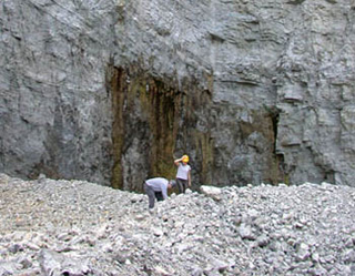 A photo of two people examining rocky ground in front of a massive cliff-face. A photo of two people examining rocky ground in front of a massive cliff-face.