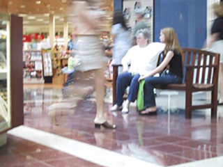 A photo of people sitting at a mall bench with people walking by. A photo of people sitting at a mall bench with people walking by.