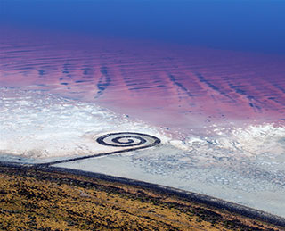 An aerial view of a spiral-shaped land art sculpture made out of mud, salt crystals, and balsalt rocks. An aerial view of a spiral-shaped land art sculpture made out of mud, salt crystals, and balsalt rocks.