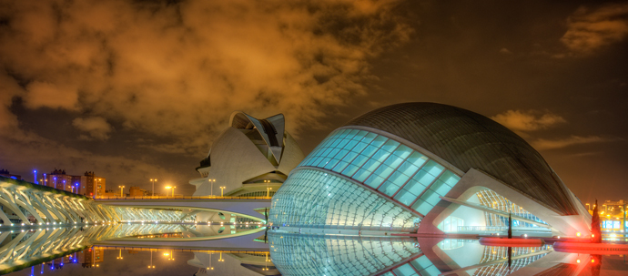 A photo of several highly stylized buildings lit at night in multiple colors, with their mirror reflection in water. A photo of several highly stylized buildings lit at night in multiple colors, with their mirror reflection in water.