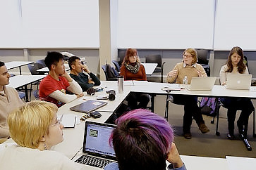 Seven students and an instructor sit around a U-shaped table. The instructor is speaking, and several students look in her direction.