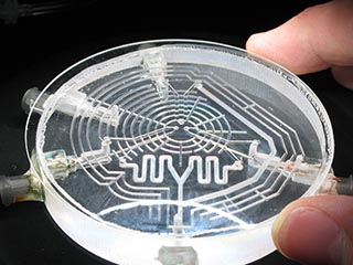 A photograph of a small, transparent, circle, shaped like a disc. It has thin lines etched into it that form patterns of concentric circles and polygons. A photograph of a small, transparent, circle, shaped like a disc. It has thin lines etched into it that form patterns of concentric circles and polygons.