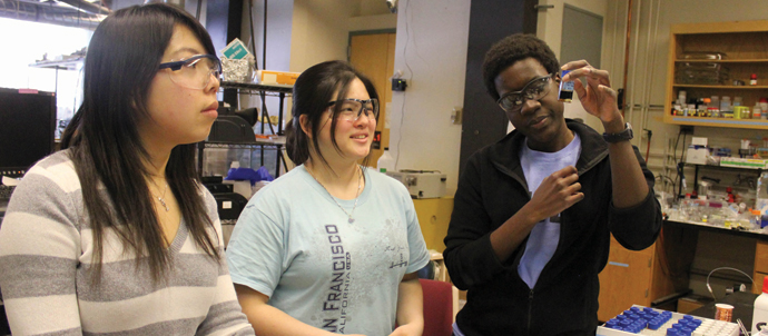 Three students in a lab examining the contents of a vial. Three students in a lab examining the contents of a vial.