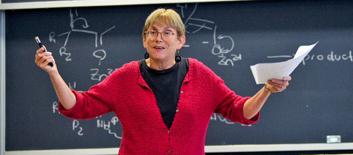 A teacher in a red sweater standing in front of a blackboard, gesturing to her audience. A teacher in a red sweater standing in front of a blackboard, gesturing to her audience.