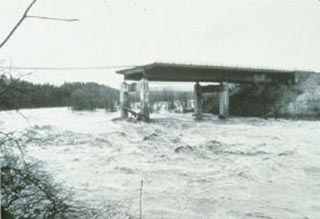 A photograph of a bridge failure in progress. Half of the bridge has been washed away and the remaning structure appears to be crumbling. A photograph of a bridge failure in progress. Half of the bridge has been washed away and the remaning structure appears to be crumbling.