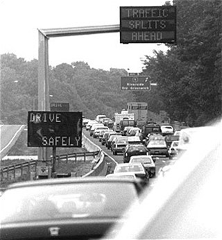 A black and white photograph of traffic congestion. A seemingly endless line of cars sit on a highway underneath signs that read 'Drive Safely' and 'Traffic Splits Ahead'. A black and white photograph of traffic congestion. A seemingly endless line of cars sit on a highway underneath signs that read 'Drive Safely' and 'Traffic Splits Ahead'.