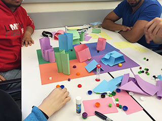 Students sitting around a homemade board game with several game pieces on it. Students sitting around a homemade board game with several game pieces on it.