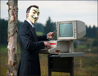 A man in a suit wears a Guy Fawkes mask on his face while standing next to a computer. A man in a suit wears a Guy Fawkes mask on his face while standing next to a computer.