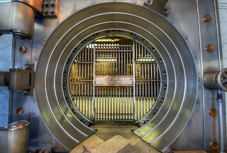 The opening of a bank vault that has metal bars surrounded by a metal circle. The opening of a bank vault that has metal bars surrounded by a metal circle.