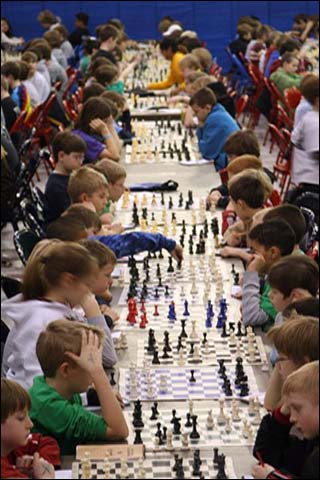 Dozens of children sit lined up at tables playing games of chess. Dozens of children sit lined up at tables playing games of chess.