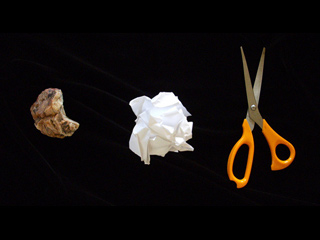 A rock, a crumpled piece of paper, and a pair of orange-handled scissors are set side-by-side on a black background. A rock, a crumpled piece of paper, and a pair of orange-handled scissors are set side-by-side on a black background.