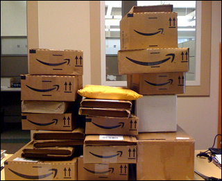 A dozen boxes from Amazon.com are stacked up on top of each other on a desk. A dozen boxes from Amazon.com are stacked up on top of each other on a desk.