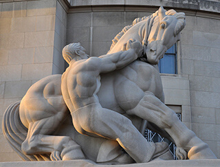 A photograph of a stone statue depicting a muscular and shirtless man pulling on the reins of an equally muscular horse. A photograph of a stone statue depicting a muscular and shirtless man pulling on the reins of an equally muscular horse.