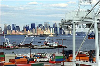 A photograph with the skyline of New York in the background and dozens of colorful shipping containers in the foreground. Ships loading and offloading float in the water. A photograph with the skyline of New York in the background and dozens of colorful shipping containers in the foreground. Ships loading and offloading float in the water.