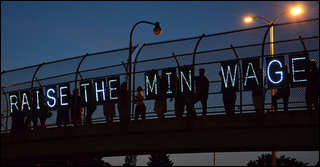 People stand on a highway overpass at night holding lighted signs that spell out "Raise the Min Wage." People stand on a highway overpass at night holding lighted signs that spell out "Raise the Min Wage."