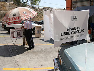 A man stands under a large umbrella, and places his votes into white boxes. A sign hangs nearby that says "el voto es libre y secreto." A man stands under a large umbrella, and places his votes into white boxes. A sign hangs nearby that says "el voto es libre y secreto."