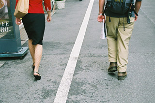 A white line divides the sidewalk. A woman walks on the left and a man on the right. A white line divides the sidewalk. A woman walks on the left and a man on the right.