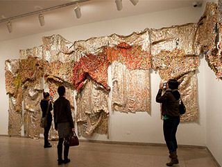 Three people stand in front of a multi-paneled drape-like sculpture made of brightly colored aluminum bottle caps and copper wire. Three people stand in front of a multi-paneled drape-like sculpture made of brightly colored aluminum bottle caps and copper wire.
