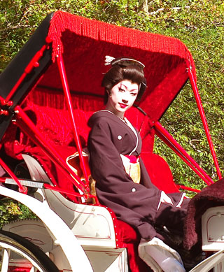A small, keen-featured man, clothed in a kimono, and wearing powder on his face and rouge on his lips, sits in a carriage. A small, keen-featured man, clothed in a kimono, and wearing powder on his face and rouge on his lips, sits in a carriage.