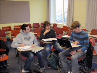 Three students sitting at desks, reading to each other from textbooks. Three students sitting at desks, reading to each other from textbooks.