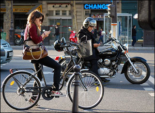 A woman on a bicycle and a man on a motorcycle are both looking down at phones in their hands while stopped in traffic. A woman on a bicycle and a man on a motorcycle are both looking down at phones in their hands while stopped in traffic.