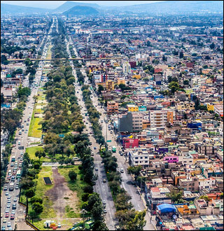 An aerial photograph showing buildings crowded together on the right side with mountains in the background. A long thin strip of green runs parallel to the roads. An aerial photograph showing buildings crowded together on the right side with mountains in the background. A long thin strip of green runs parallel to the roads.