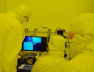People in cleanroom suits view magnified wafer pattern on screen. People in cleanroom suits view magnified wafer pattern on screen.