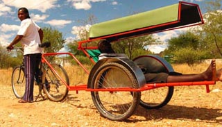 Photo of a two-wheeled ambulance cart attached to rear of bicycle, carrying a reclining patient. Photo of a two-wheeled ambulance cart attached to rear of bicycle, carrying a reclining patient.