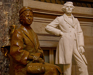 A bronze statue of a seated woman with her hands folded close to her purse is positioned near the marble statue of a standing gentleman wearing a knee-length coat, with one hand on his hip. A bronze statue of a seated woman with her hands folded close to her purse is positioned near the marble statue of a standing gentleman wearing a knee-length coat, with one hand on his hip.