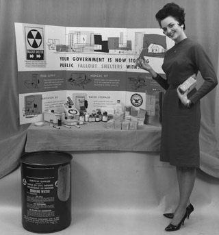 An attractive woman stands in front of a display featuring individually-sized supplies for a fallout shelter, such as food, and medical and sanitation kits. An attractive woman stands in front of a display featuring individually-sized supplies for a fallout shelter, such as food, and medical and sanitation kits.