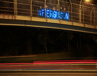 Protesters stand on a highway overpass with a lit up sign that reads "#FERGUSON". Protesters stand on a highway overpass with a lit up sign that reads "#FERGUSON".