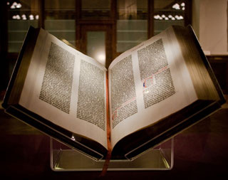 A large book, placed on a Plexiglas stand, lies open to a page containing elaborate type. A large book, placed on a Plexiglas stand, lies open to a page containing elaborate type.