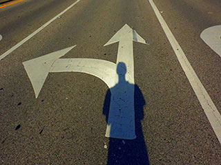 A man's shadow falls on two arrows painted on a road—one arrow points left and the other points straight ahead. A man's shadow falls on two arrows painted on a road—one arrow points left and the other points straight ahead.