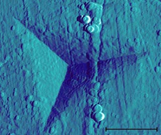 A sharp probe has left a triangular indentation in a surface, shown using scanning electron microscopy. A sharp probe has left a triangular indentation in a surface, shown using scanning electron microscopy.