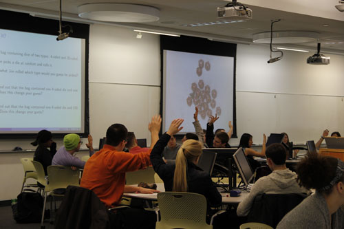 <p>In many class meetings, we illustrated concepts using physical objects like dice and cards. Sometimes, this took the form of an instructor-led demonstration in front of all of the students. We often asked students to participate by choosing cards, guessing outcomes, and explaining their ideas.</p> <p>At the start of the course, we also gave each student a bag of dice that included 10-sided dice and all the Platonic solids. The students used the dice to explore probability and collect data for statistics.</p> <p>In the demonstration pictured here, we illustrated the role of prior probability in Bayesian inference. We told students that we would randomly select and roll a die from a cup containing dice with 4-sides and 20-sides. Secretly the cup contained twenty 20-sided dice and one 4-sided die, and we intentionally placed the 4-sided die on top of the pile in order to select it by feel. We rolled the die covertly, reported a 3, and asked students which type of die was most likely chosen; students were split between 4-sided and equally likely. We then used the document camera to reveal the contents of the cup; as shown, the students now overwhelmingly favored the 20-sided die. This demonstration can be seen in the sample class video.</p> Students seated at round tables raise their hands. One projector screen shows a slide and one projector screen shows a set of dice.