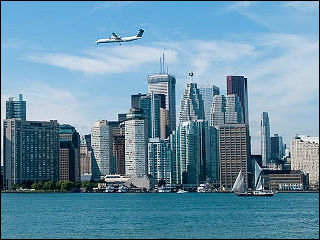 Photograph of the Toronto skyline seen from over the water, with a commercial jet in flight over the skyline and boats visible near the harbor. Photograph of the Toronto skyline seen from over the water, with a commercial jet in flight over the skyline and boats visible near the harbor.
