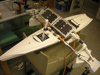 A long slim white hull sits in a cradle, with PVC/Styrofoam pontoons extending to the sides, and a solar panel and periscope assembly sitting on top. A long slim white hull sits in a cradle, with PVC/Styrofoam pontoons extending to the sides, and a solar panel and periscope assembly sitting on top.