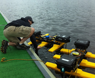 A man crouched on a river dock, reaching down to two small floating “robots” that consist of several boxes of electronics supported on pontoons. A man crouched on a river dock, reaching down to two small floating “robots” that consist of several boxes of electronics supported on pontoons.