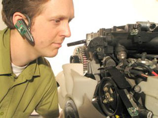 A man wearing a bluetooth earset is crouched next to an internal combustion engine. A man wearing a bluetooth earset is crouched next to an internal combustion engine.