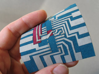 Photo of a hand holding a small fabric square with a "printed circuit" pattern of connectors on it. Photo of a hand holding a small fabric square with a "printed circuit" pattern of connectors on it.